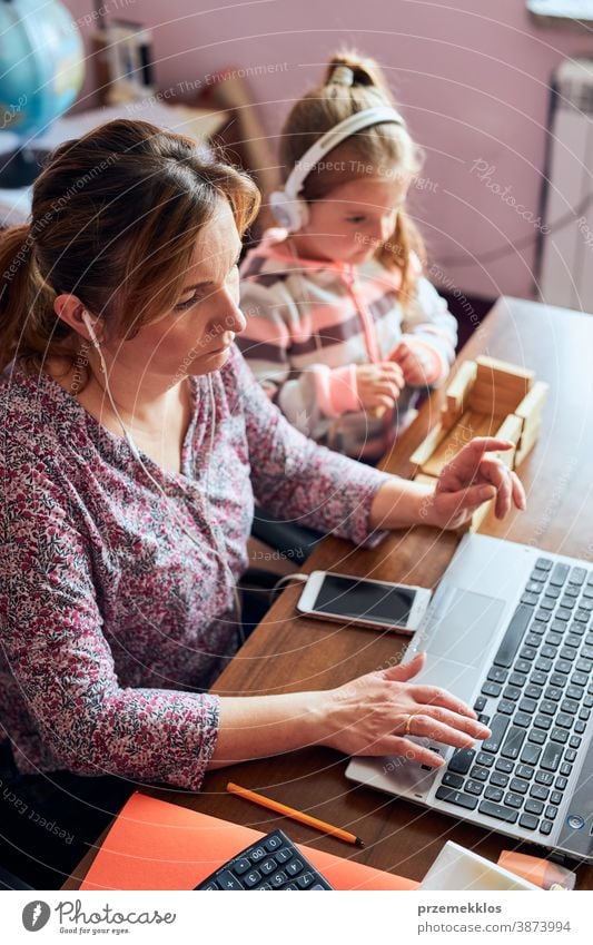 Woman mother working doing her job remotely during video chat call stream online course webinar on laptop from home while her daughter playing with bricks toy