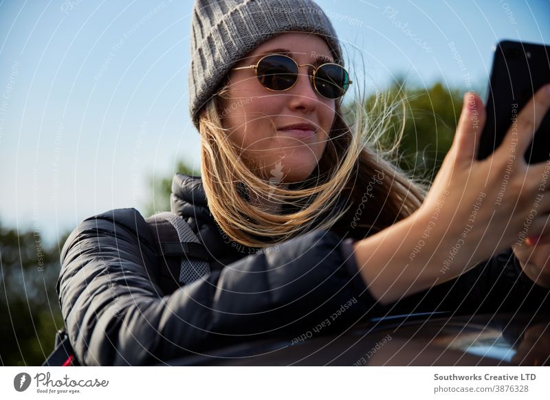 Young Woman On Road Trip Vacation Leaning On Roof Of Rental Car Using Mobile Phone social media cell phone road trip woman young women holiday vacation car