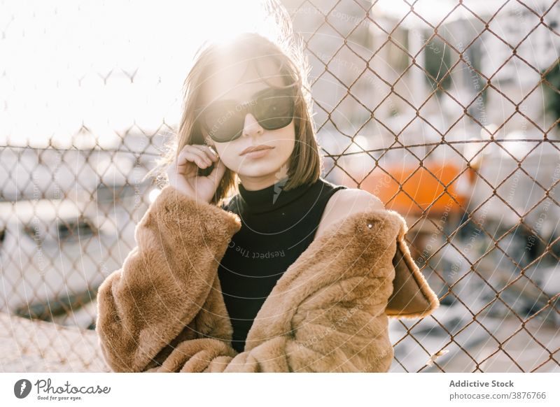 Stylish woman in warm jacket in city fur style trendy outfit determine sunglasses outerwear female net fence street urban cool fashion serious town calm