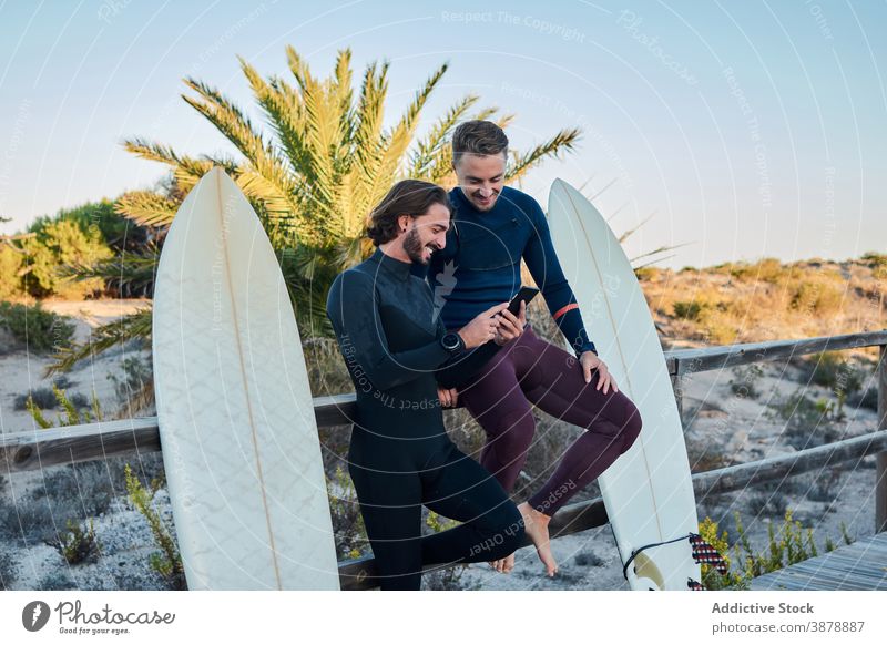 Cheerful surfers using smartphone together on embankment promenade surfboard seaside cheerful gadget boardwalk device summer friend friendship browsing