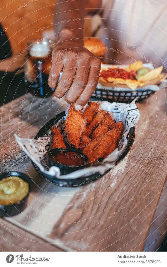 Crop Black man eating fast food in cafe chicken wing ethnic wooden delicious junk food black enjoy sauce yummy lunch tasty male fresh table sit appetizing