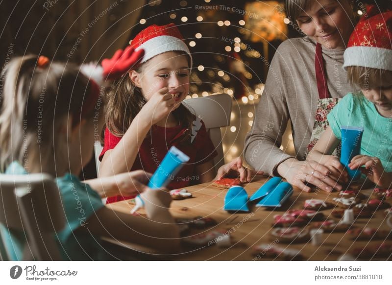 Mother and little kids in red hats cooking gingerbread cookies and decorating with glaze. Beautiful living room with lights and Christmas tree. Happy family celebrating holiday together.