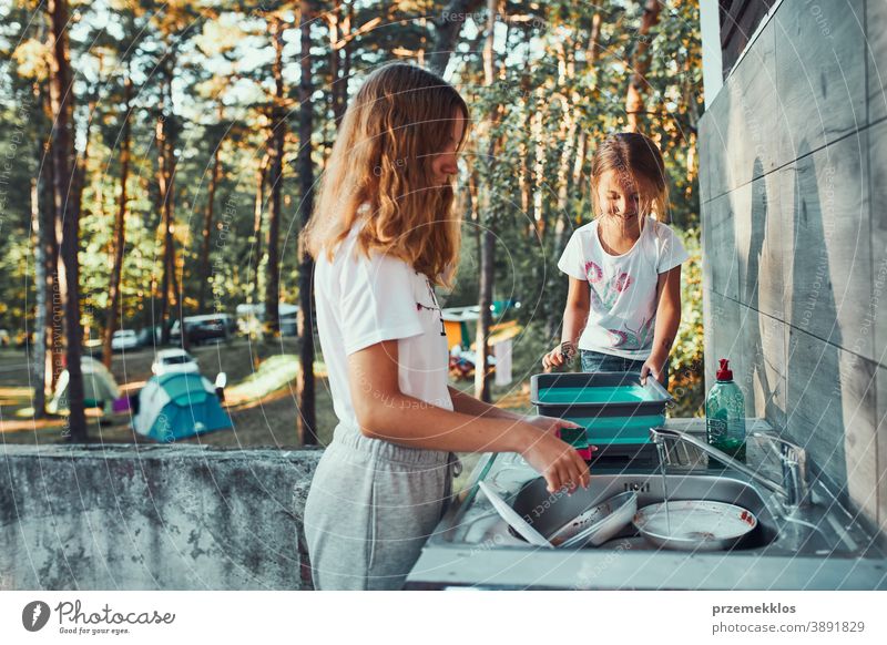 Teenager girl washing up the dishes pots and plates with help her younger sister in the outdoor kitchen during vacations on camping working together siblings