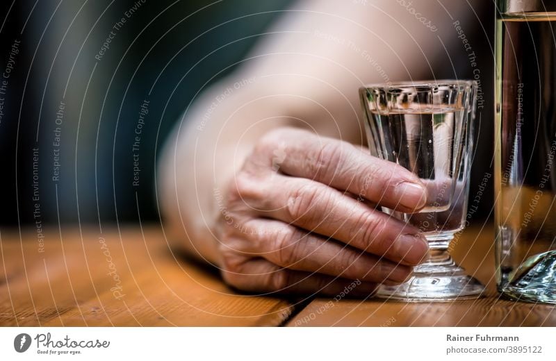 On a table there is a bottle of alcohol and a shot glass. A person's hand is holding the shot glass. Anonymous Addiction addicted Alcoholic drinks booze drunken