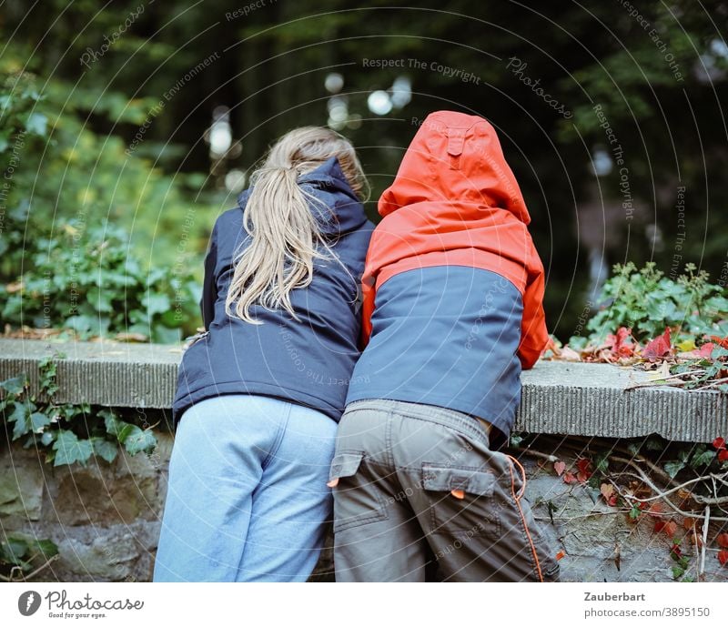 Two children in anoraks lean over a wall and look down Boy (child) Girl Anorak Rain jacket Stand Wall (barrier) Interest Trust Curiosity Red Blue Green mystery