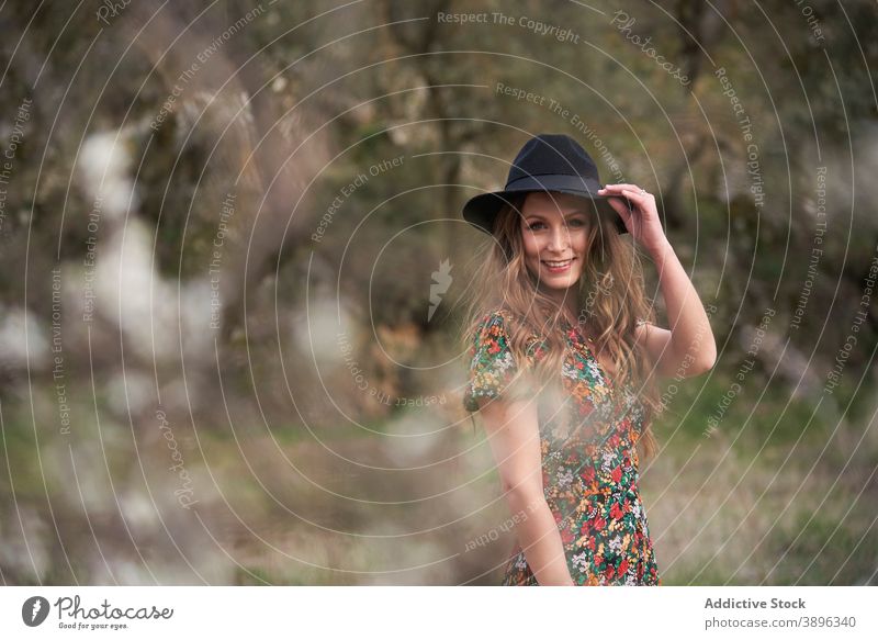 Glad woman in hat standing in forest accessory charming dress smile outfit nature apparel female woods appearance long hair wavy hair optimist style positive