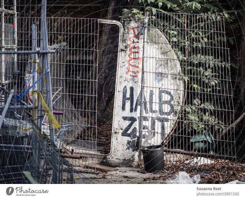 A broken building fence, in front and behind it building rubble, painted with graffiti. With black paint the word half time is written on a semicircular construction element.