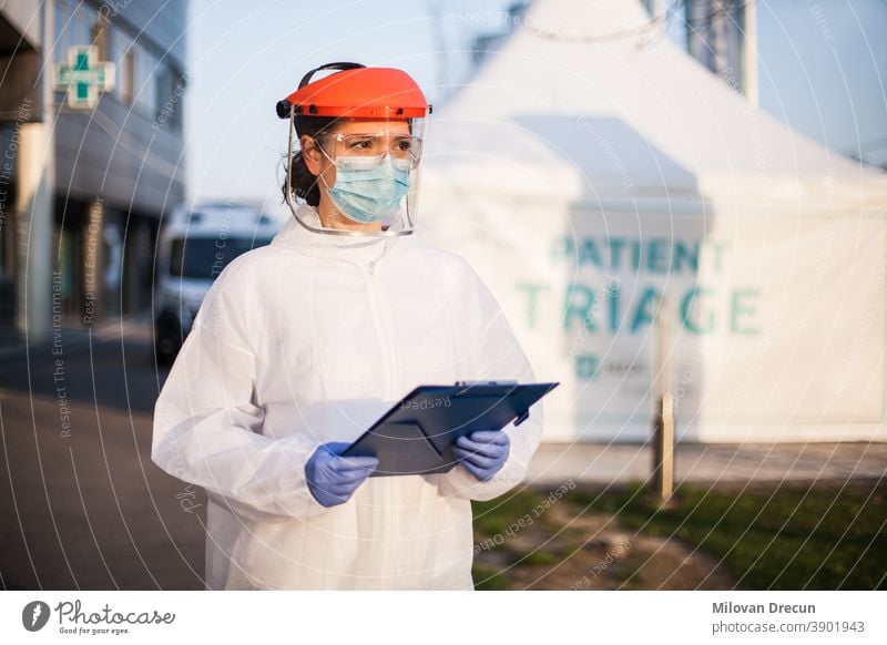 Paramedic wearing personal protective equipment PPE holding folder standing in front of ICU hospital isolation rt-PCR drive thru testing site,COVID-19 pandemic outbreak crisis,worried exhausted staff