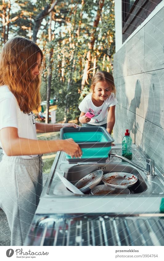 Teenager girl washing up the dishes pots and plates with help her younger sister in the outdoor kitchen during vacations on camping working together siblings