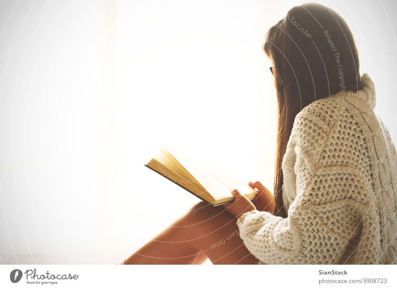 Young woman sitting in bed while reading a book window view lazy sunday winter coffee cup tea glasses morning girl home relax socks beautiful young bedroom