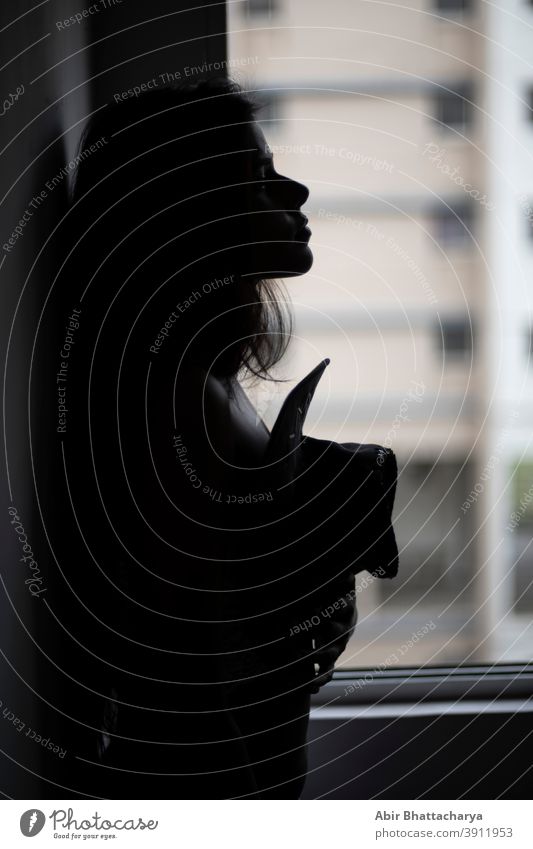 Silhouette portrait of a young and attractive dark skinned Indian Bengali woman in lingerie with a hat posing in a casual mood in front of a window. Boudoir photography.