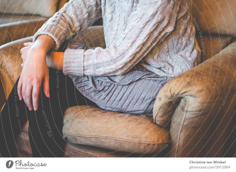 A young woman sits slouched in a faded brown leather chair with her legs crossed. She wears a grey cable-knit sweater, a grey pleated skirt and black stockings. Portrait without head