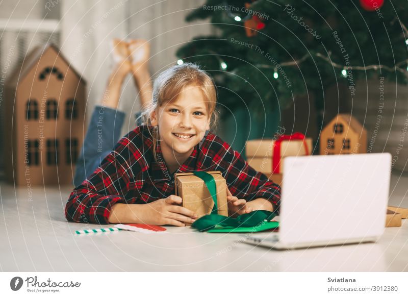 A happy little girl opens a gift under the Christmas tree at home and communicates with her grandparents via a laptop via video link. Holiday concept for Christmas and New Year at home.