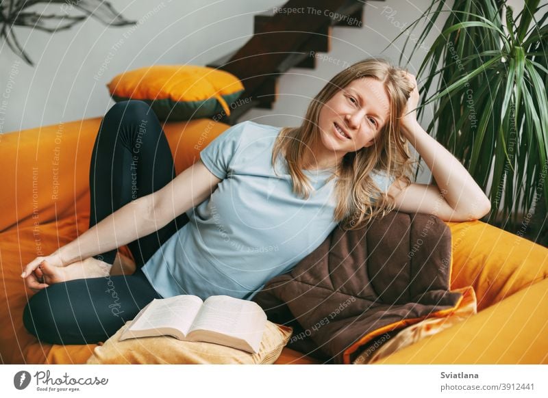 A young woman is sitting on a comfortable sofa in the room and smiling, with an open book next to her. The girl ponders what she has read in the book. home