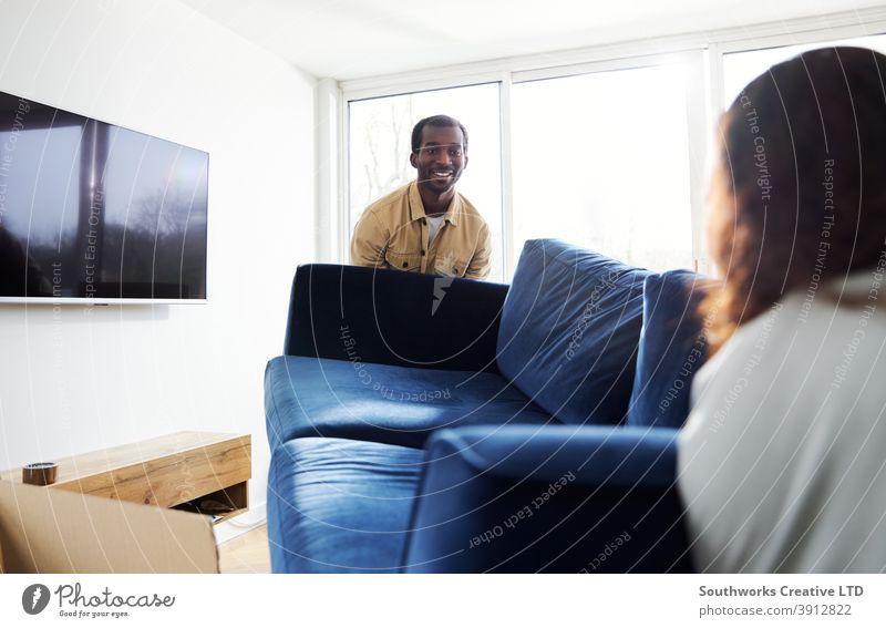 Young Couple In New Home Carrying Sofa In Lounge On Moving Day Surrounded by Removal Boxes couple young couple house buying carrying lifting furniture sofa