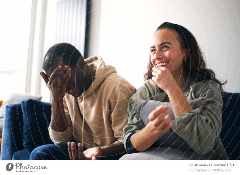 Smiling Young Couple Relaxing At Home Sitting On Sofa Cringing Whilst Watching Film On TV Together couple young couple at home sitting sofa lounge watching tv