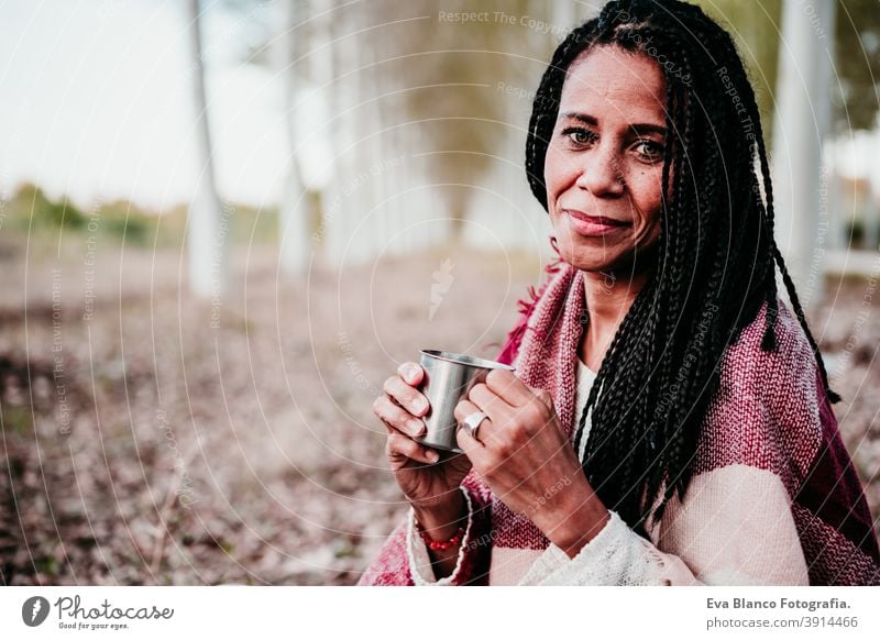 portrait of hispanic mid adult woman outdoors holding wrapped in blanket. Holding mug of water water during picnic.Autumn season autumn afro woman latin sunset