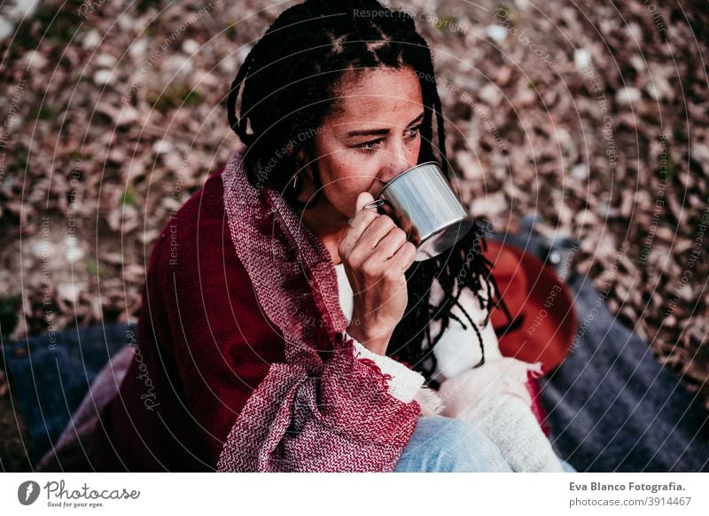 portrait of hispanic mid adult woman outdoors holding wrapped in blanket. drinking water during picnic.Autumn season autumn afro woman latin sunset nature hat