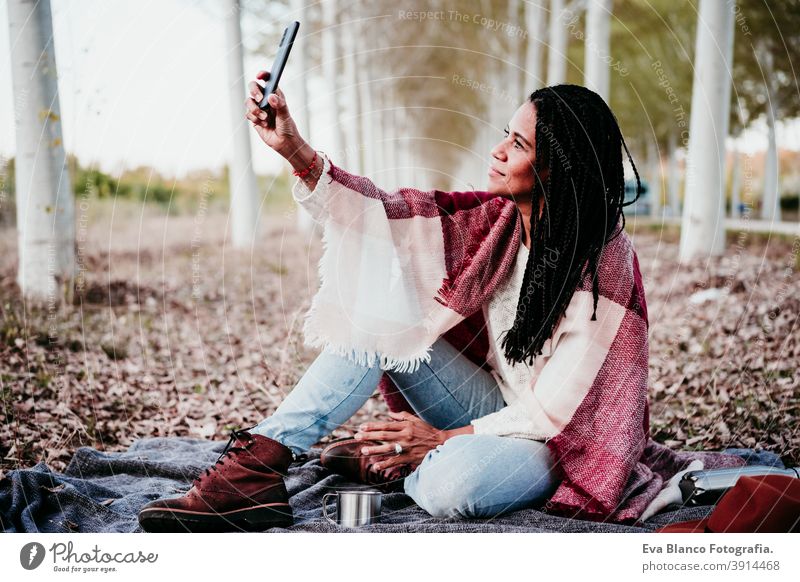 portrait of hispanic mid adult woman outdoors holding wrapped in blanket. Using mobile phone during picnic.Autumn season autumn afro woman latin sunset nature