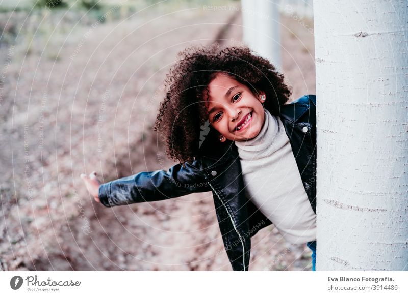 portrait of cute afro kid girl hiding behind a tree outdoors. Childhood and happiness fun autumn nature golden hour hat brown trees leaves casual clothing happy