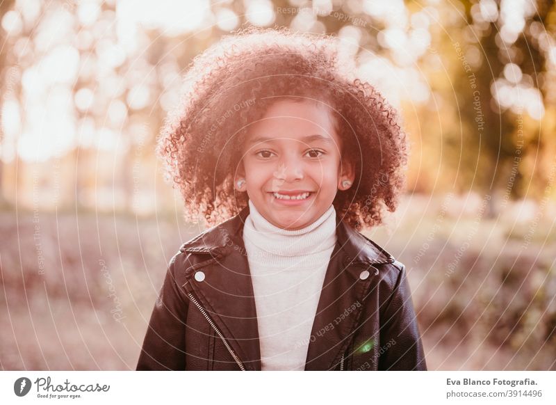 portrait of cute afro kid girl smiling at sunset during golden hour, autumn season, beautiful trees background nature outdoors hat brown leaves casual clothing