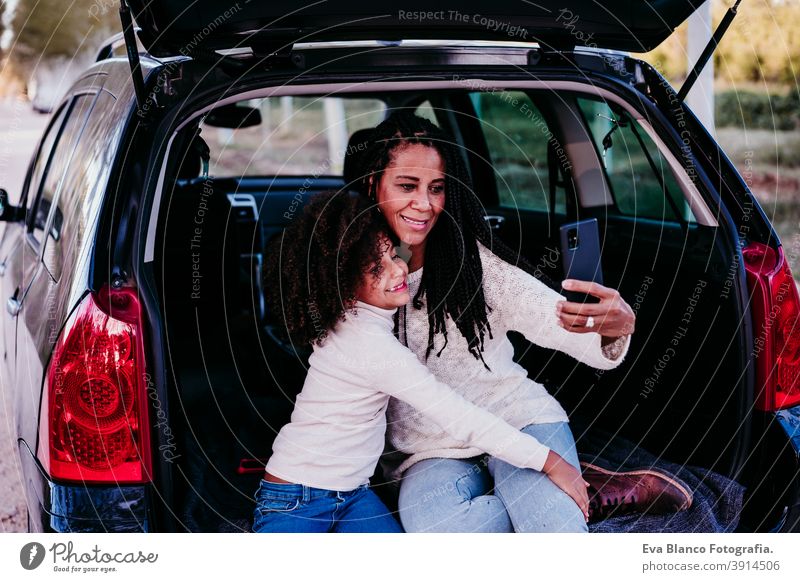 hispanic mother and afro kid girl sitting in a car in nature. Having fun and taking pictures with mobile phone. Autumn season. Family and travel concept