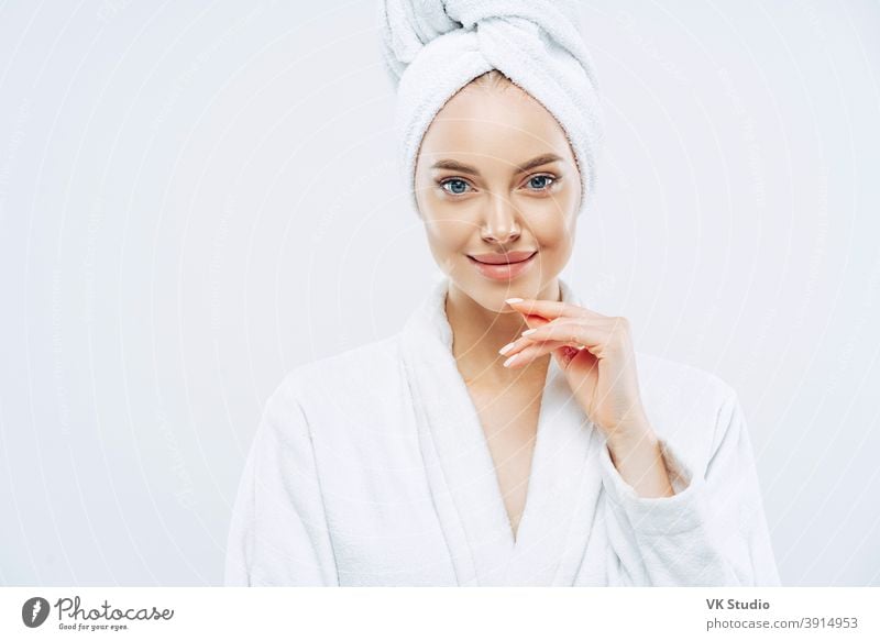 Studio shot of pretty beauty woman has washed hair, wears wrapped towel on head, has manicure, cute natural face, touches chin gently, looks with tender smile, dressed in bath robe, poses indoor