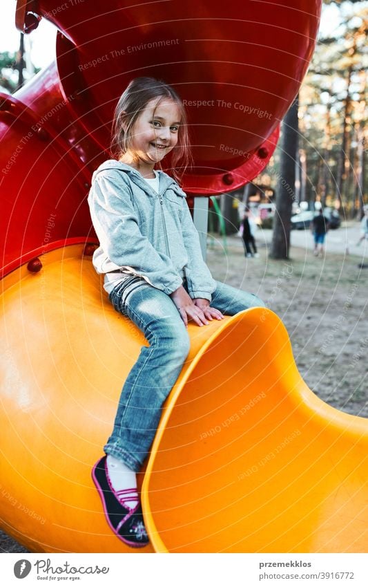 Little girl preschooler playing on a playground sitting on tube slide smiling and looking at camera positive joyful junior public place nursery climb outdoors