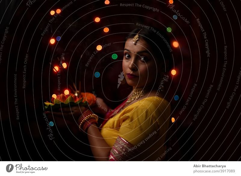 An young and beautiful Indian Bengali woman in Indian traditional dress is holding a Diwali diya/lamp in her hand in front of colorful bokeh lights. Indian lifestyle and Diwali celebration