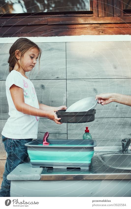 Teenager girl washing up the dishes pots and plates with help her younger sister in the outdoor kitchen during vacations on camping working together siblings