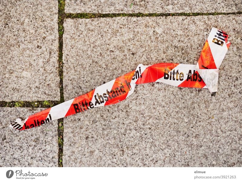 red and white barrier tape with the inscription "Please keep your distance" lies crumpled on the grey concrete pavement / Corona keep sb./sth. apart