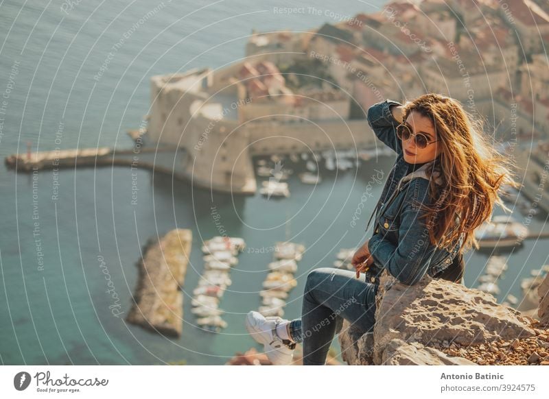 Attractive brunette seen from behind posing while sitting on a stone above the city of Dubrovnik, outlines of the majestic old town in the background. Traveler visiting the walls and the Srd mountain