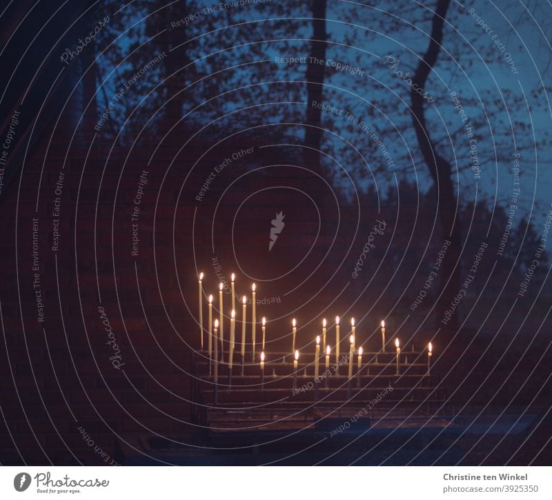 Candle altar with burning candles in a chapel, photographed through a glass door. It reflects trees and the evening sky at blue hour Sacrificial candles