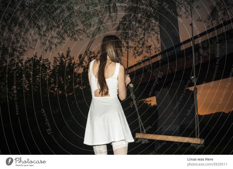 A gorgeous brunette girl dressed in a white dress under the bridge. It’s just after the sunset and the sky is all orange and purple. Pure romanticism in the city park.