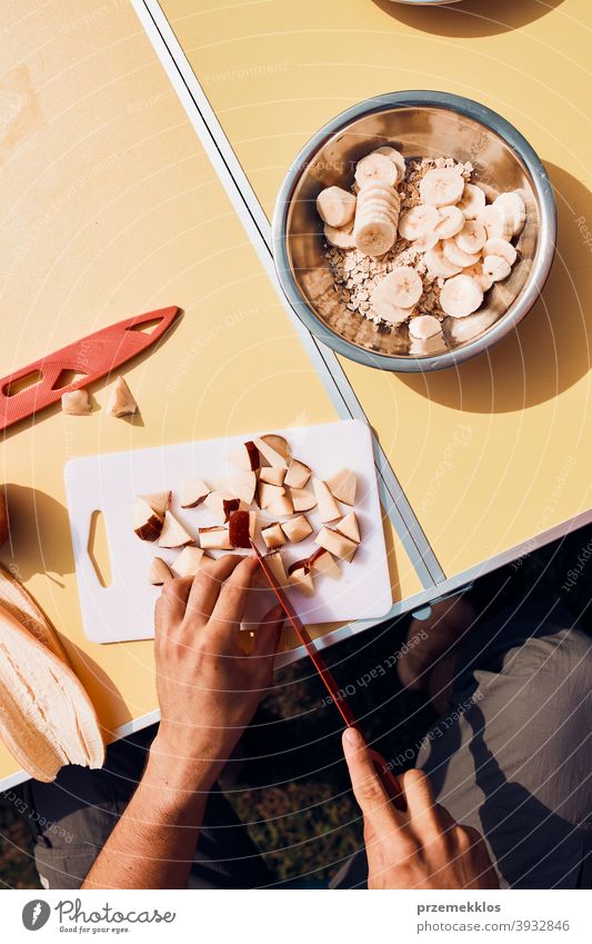 Man preparing healthy breakfast with fruits and oat flakes outdoors on camping. Close up of male hands cutting apples and bananas for breakfast table board