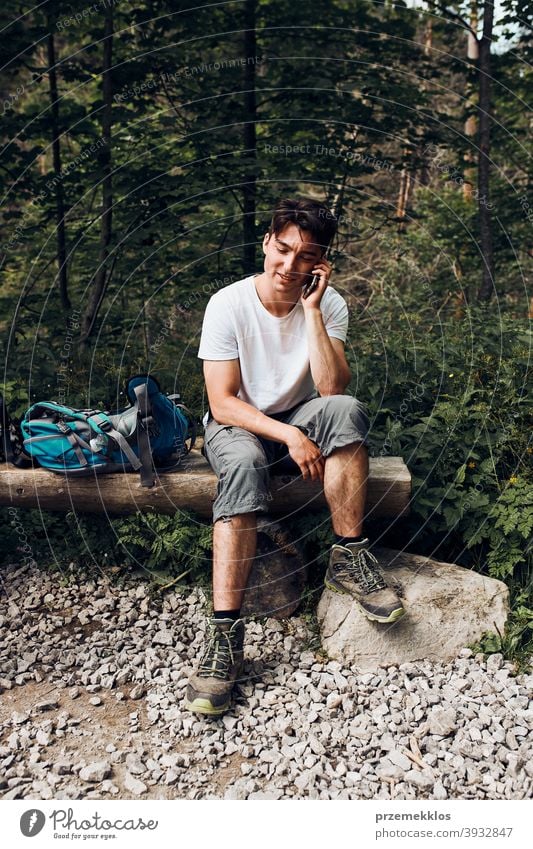 Young man with backpack talking on phone having break sitting on trunk during hike in a mountains activity adventure forest forest landscape forest path freedom