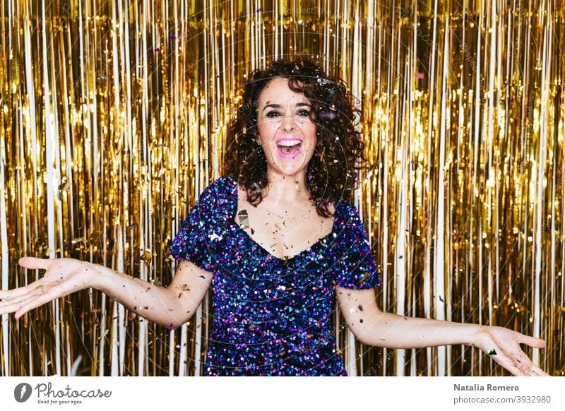 A beautiful woman in elegant clothes with Christmas decorations behind her celebrating New Year's Eve while some confetti is falling above her. New Year's Eve party at home concept