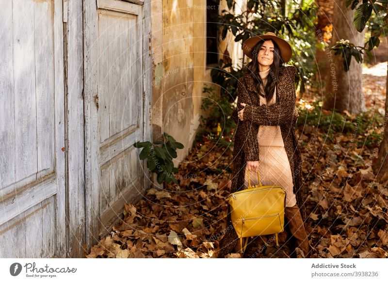 Stylish young ethnic woman standing in autumn garden near shabby house park fashion door smile trendy foliage leaf female long hair brunette style outfit hat