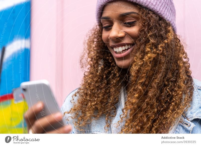 Happy young ethnic lady in stylish outfit browsing smartphone near pink door woman read message smile cheerful trendy street joy mobile female teenage