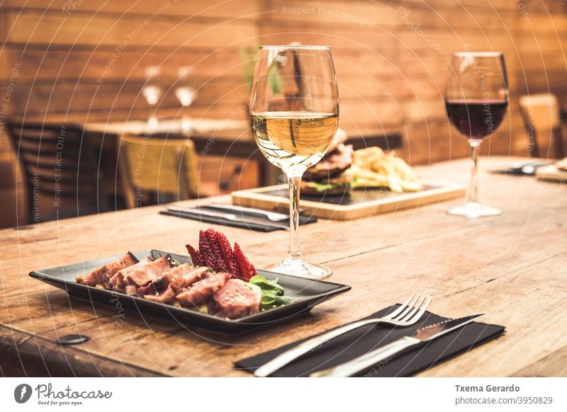 Traditional restaurant atmosphere with tuna tataki and burger with chips in the background on rustic wooden table and glass of white wine fish tomato cup