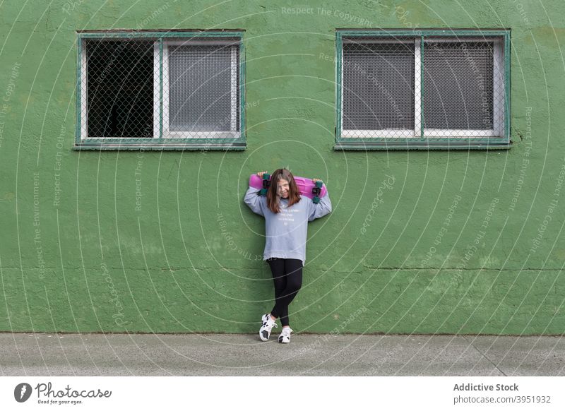 Positive stylish female teenager resting near green building with skateboard in hands girl relax training activity hobby break holiday style street smile young