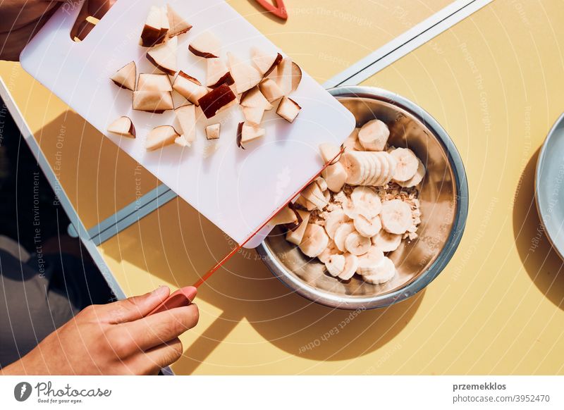 Man preparing healthy breakfast with fruits and oat flakes outdoors on camping. Close up of male hands cutting apples and bananas for breakfast table board