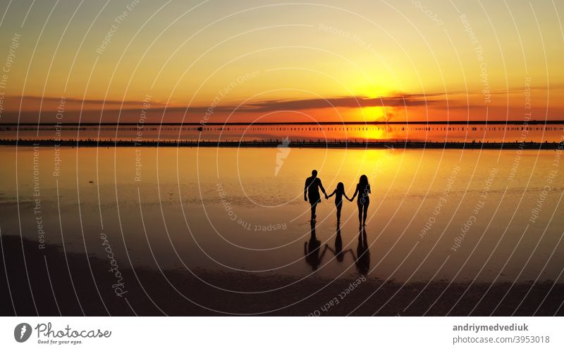 Silhouettes of three, dad, mom and little daughter walking through the shallows of a lake against the background of a red-orange sunset two silhouettes people