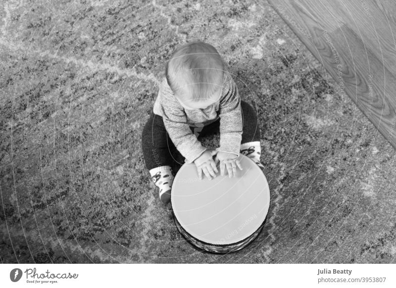 6 month old baby playing drum while seated on a rug on the floor; black and white image from bird's eye view floor drum 6 months old hands beat tap scratch