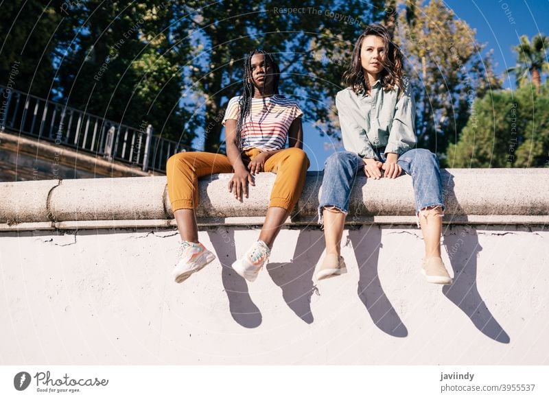 Two multiethnic women sitting on a urban wall. serious expression sunlight friend black afro girl student two people lifestyle female pretty young friendship