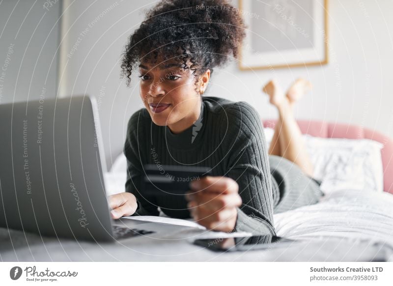 Young woman lying on bed at home with laptop computer using credit card to shop online bedroom debit card banking pay day tech technology connected the internet