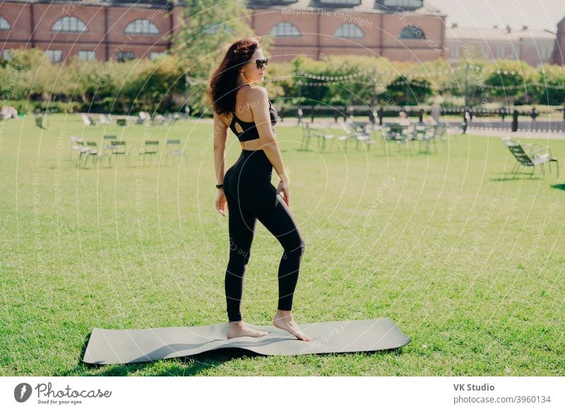 Fit brunette woman dressed in sportswear stands bare feet on karemat has fintess training on green lawn outdoor during summer time wears sunglasses looks somewhere being in good physical shape