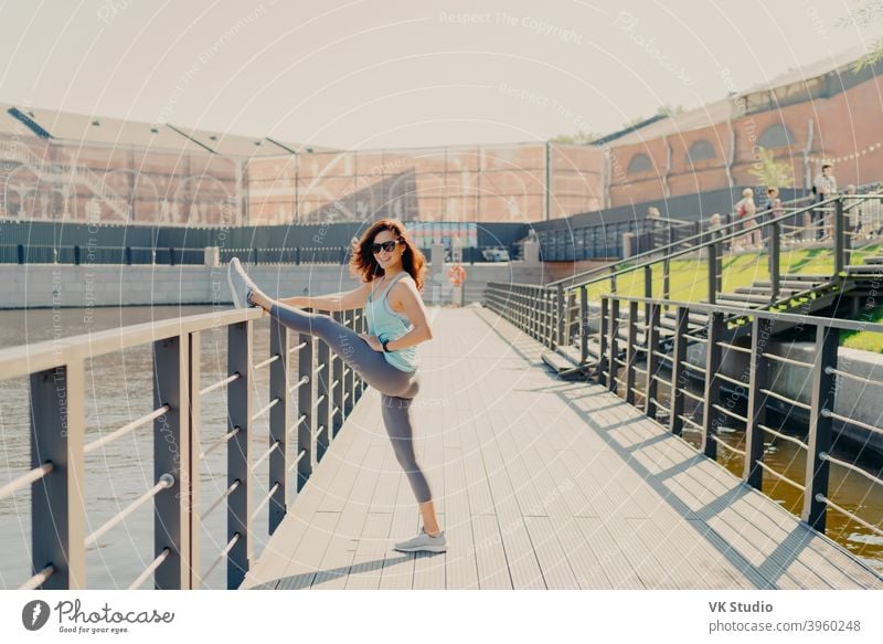 Glad brunette woman leans at fence stretches legs has perfect figure dressed in activewear poses outdoor demonstrates her flexibility leads sporty lifestyle warms up before fitness training.