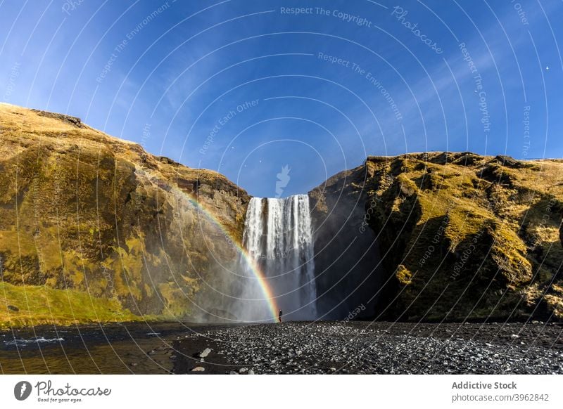 Explorer standing near waterfall on sunny day rainbow traveler mountain vacation landscape amazing flow stream tourism iceland explore explorer nature