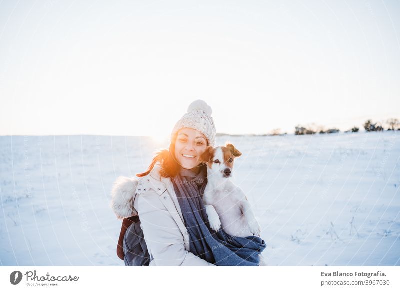 happy woman in snowy mountain wearing modern coat at sunset. Holding cute jack russell dog in arms. winter season. nature at sunset travel owner pet love